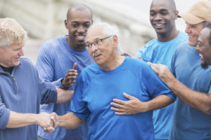 Multi-ethnic group of men wearing blue shirts outdoors, congratulating their friend, a senior Hispanic man in his late 70s.